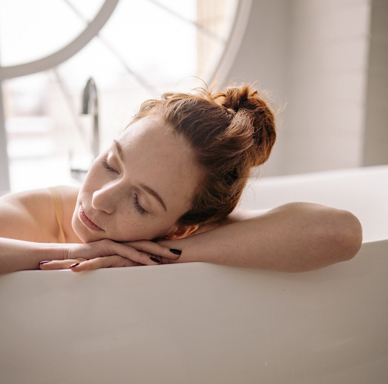 A woman sleeping with her head on her hands on the side of the tub.