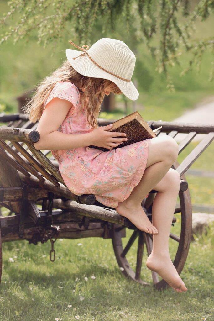 a girl wearing a pink dress sitting on a park bench wearing a straw hat reading a book from a list of health and wellness books