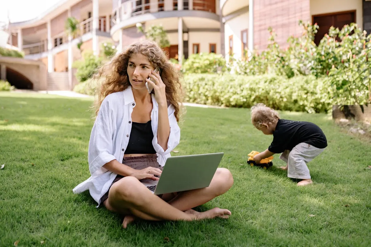 a woman outside on her cell phone and laptop, babysitting her toddler